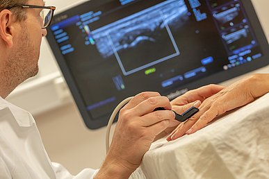 Arzt fährt mit einem fingerdicken Ultraschallgerät über einen Finger einer ausgestreckten Hand. Auf einem dahinter befindlichen Monitor ist blau das Ergebnis der Durchleuchtung zu sehen.