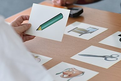 Auf einen Tisch liegen weiße Karten mit jeweils einer Zeichnung darauf: Bett, Schere und Maske. Eine Person, von der nur die Hand zu sehen ist hält eine Karte mit der Abbildung eines Bleistifts.