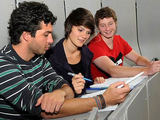 3 junge Erwachsene betrachten im Hörsaal gemeinsam ihre Unterlagen.