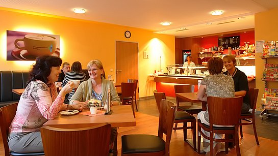 Einige unterhalten sich entspannt bei Kaffee und Kuchen in der in warmen Orangetönen gehaltenen Cafeteria. 