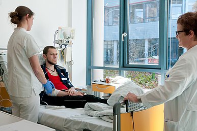 Ein Patient sitzt auf einem Bett, während eine Mitarbeiterin mit blauen Handschuhen, über einen Zugang eine Spritze gibt. Der Patient blicht zu einer weiteren Mitarbeiterin in weißer Klinikkleidung, die am Bettende steht.