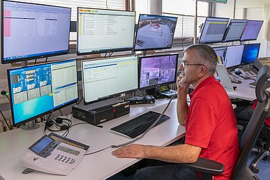 Mann im roten T-Shirt, mit grauen Haaren und einer Brille blickt konzertiert  in einen von 12 Monitoren eines Kontrollraums während er ein Telefongespräch führt.