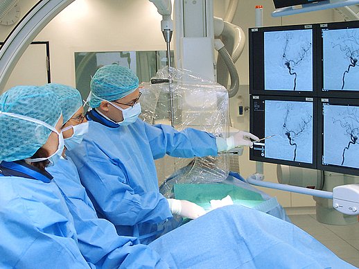 3 Mediziner stehen links eines abgedeckten Patienten im OP-Saal und betrachten 4 Bildschirme mit Aufnahmen von Gefäßen mit Kontrastmitteln..