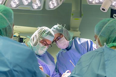 In einem Operationssaal blicken zwei Operateure konzentriert auf ihren Patienten, der selbst nicht zu sehen ist. Oben und seitlich ist der Blick auf die beiden Personen durch eine OP-Lampe und zwei Kollegen in Rückenansicht eingerahmt.