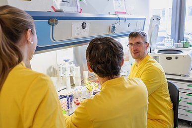Drei Labormitarbeiter in gelben Laborkitteln sitzen und stehen diskutierend an einer Laborbank mit verschiedenen Reagenzien.