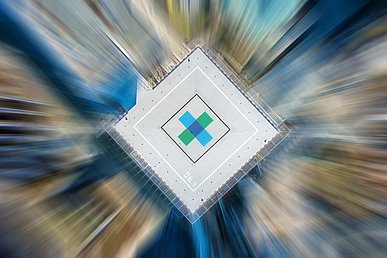 Aufsicht auf den Hubschrauberplatz. In die Landezone ist das Logokreuz des Uniklinikums Erlangen in den Farben Dunkelblau, Hellblau und Grün montiert. Durch einen Filtereffekt werden die Konturen umliegenden Gebäude in unscharfe stürzende Linien verwandelt, was einen Bewegungseffekt erzeugt.
