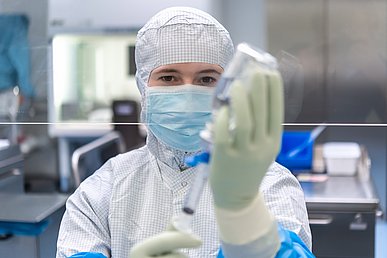 Labormitarbeiterin in geschlossener Schutzkleidung zieht eine Spritze mit einen klaren Flüssigkeit aus einer Glasflasche auf.