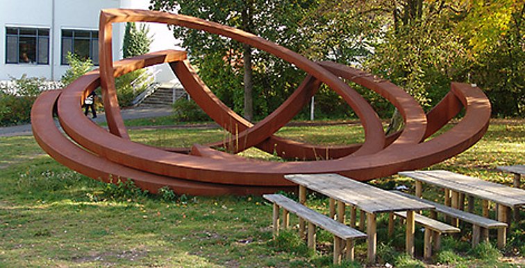 Das Kunstwerk besteht aus drei ca. 6 Meter breiten bronzefarbenen Metall-Ringen, die an einer Seite wie zusammengefaltet abknicken.