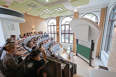 Blick von oben in einen modern renovierten historischen Hörsaal. Die im Viertelkreis angeordneten Sitzreihen sind mit Studierenden gut gefüllt. 
