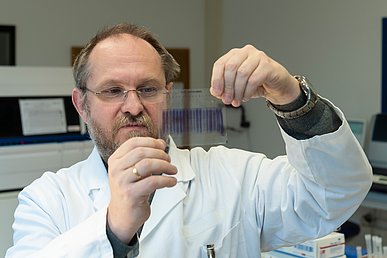 Wissenschaftler in weißem Laborkittel hält mit beiden Händen eine durchsichtige Folie empor und blickt konzentriert auf die darauf befindlichen blauen Marker.