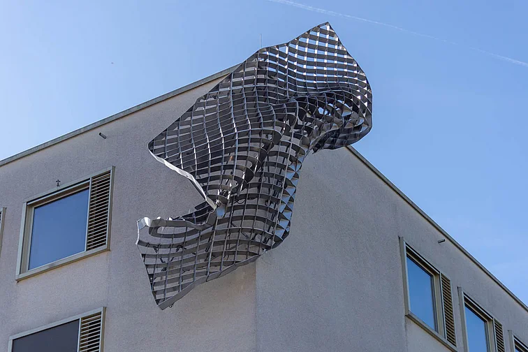 An der Hausecke kurz unterhalb des Daches ist eine mehrere Quadratmeter große Metallskulptur befestigt. Sie ist geformt wie ein in sich verdrehtes Metallgitter.