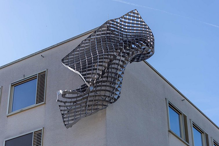 An der Hausecke kurz unterhalb des Daches ist eine mehrere Quadratmeter große Metallskulptur befestigt. Sie ist geformt wie ein in sich verdrehtes Metallgitter.