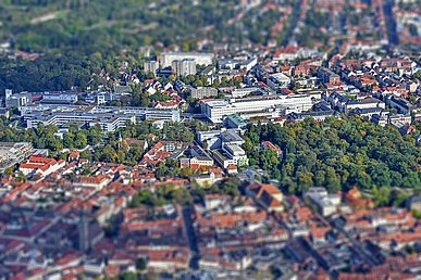 Luftaufnahme von West nach Ost. Die über die gesamte Breite quer liegenden Klinikgebäude des Internistischen und Operativen Zentrums sind scharf abgebildet. Die roten Hausdächer im Vordergrund und der Bildhintergrund sind unscharf.