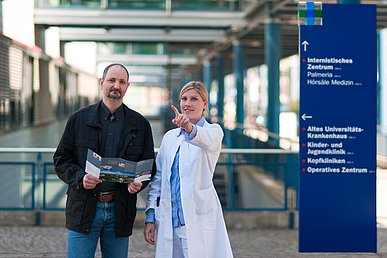 Frau in weißem Kittel weist mit der Hand bärtigem Mann mit schwarzer Jacke den Weg. Rechts eine dunkelblaue Wegweiser-Tafel mit weißer Beschriftung und dem Logo des Uniklinikums Erlangen. Unscharf im Hintergrund der Außenbereich eines modernen Klinikgebäudes.