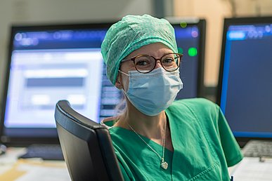 Ärztin in grüner Funktionskleidung mit OP-Haube, Brille und Schutzmaske hat sich auf einem Schreibtischstuhl zur Kamera gedreht. Im Hintergrund sind unscharf mehrere Überwachungsmonitore zu sehen.