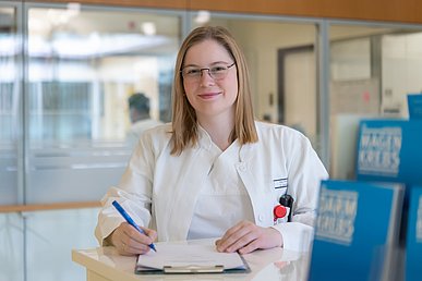 Junge Frau in weißer Klinikkleidung steht an einem Tresen, auf dem ein Klemmbrett liegt, mit einem Stift in der Hand und lächelt in die Kamera.