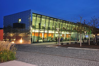 Nachtansicht der erleuchteten, zweigeschossigen Glasfront der Hörsäle Medizin mit Blick in das Foyer und dem Gang zu den Seminarräumen im Obergeschoss.