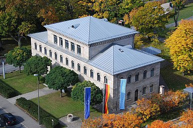 Die dreigeschossige Sandsteinvilla der Kaufmännischen Direktion mit grauem Zeltdach aus Blech fotografiert von schräg oben umrahmt von buntem Herbstlaub. Im Vordergrund die europäische die deutsche und die bayerische Fahne.