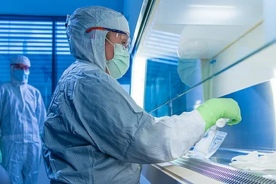 Labormitarbeiterin in geschlossener Schutzkleidung arbeitet an einer Laborbank. Schräg hinter ihr steht eine weiterer Mitarbeiter in Schutzkleidung und sieht zu.