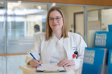 Aufklärungsarbeit: Biomaterial für die Forschung freigeben Junge Frau in weißer Klinikkleidung steht an einem Tresen, auf dem ein Klemmbrett liegt, mit einem Stift in der Hand und lächelt in die Kamera.