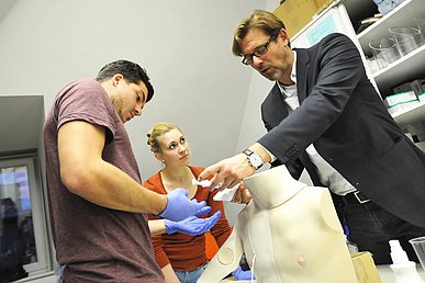 Dozent zeigt zwei Studierenden an einer Simulationspuppe die richtige Handhabung einer Spritze.