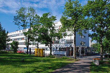 Der langestreckte viergeschossige Ostflügel der Kinder und Jugendklinik im Sonnenlicht, im Vordergrund die Grünanlage des Maximiliansplatzes.