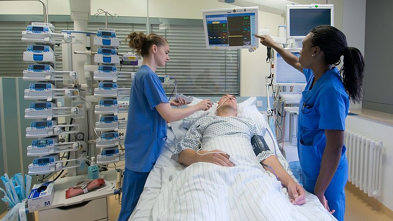 Zwei Mitarbeiterinnen in blauen Kasacks betreuen einen Patienten mit Nasensonde umgeben von Apparaten zur Medikamentengabe und Monitoren mit Vitalfunktionen.