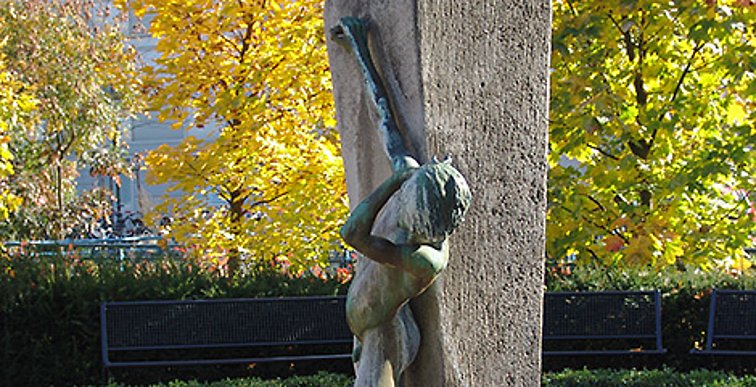 Grün angelaufene Figur eines nackten Jungen der seinen rechten Arm nach oben streckt und ein Rohr zuhält, während sein linker Arm an die eigene Schulter fasst.