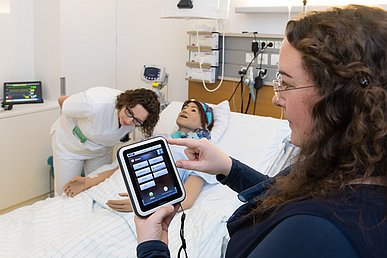 Auf einem Patientenbett liegt eine Simulationspuppe. Eine Pflegeschülerin hört die Puppe ab. Im Bildvordergrund ist eine Ausbilderin zu sehen, die über ein Steuergerät verschiedene Aktivitäten bei der Puppe simulieren kann.