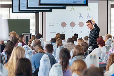 Rückenansicht einer Gruppe Studierenden, die in einem Seminarraum auf eine Projektion über Blutgruppen blicken. In der Mitte des Raumes steht ein Dozent vor einem Mikroskop, der zwei Röhren empor hält und zu den Studenten blickt.