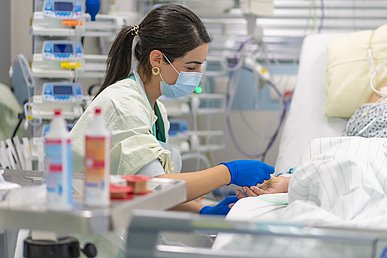 Intensivpflegerin legt einem Patienten am Handgelenk einen Zugang. Im Hintergrund sind Spritzenpumpen und weiteres Equipment eine Intensivstation zu sehen.
