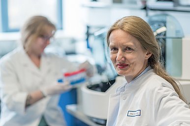 Endokrinologische Forschung in der Medizinischen Klinik 1 – Gastroenterologie, Pneumologie und Endokrinologie Porträtaufnahme einer Wissenschaftlerin in weißer Laborkleidung. Unscharf abgebildet bestückt im Hintergrund bestückt eine Labormitarbeiterin eine Zentrifuge.