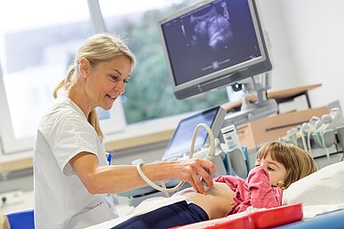 Ärztin in weißer Funktionskleidung untersucht ein Mädchen mit einem Ultraschallgerät. Das Untersuchungsbild des Bauchs ist auf einem Monitor zu sehen.