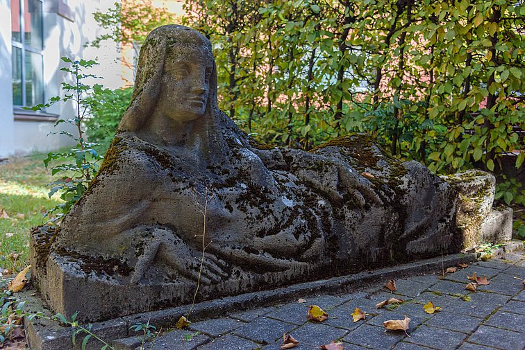 Steinfigur einer entspannt seitlich auf den Arm gestützt liegenden Frau.