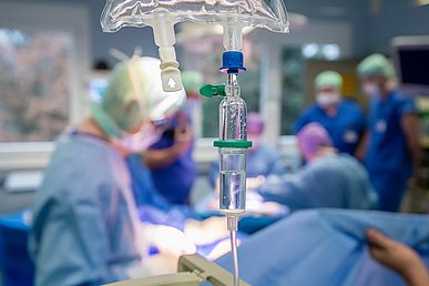 Scharf abgebildet ein Tropfer, der an einen Infusionsbeutel hängt. Unscharf im Hintergrund mehrere Personen in blauer OP-Kleidung am OP-Tisch während einer Operation.