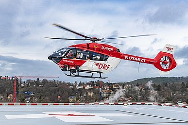 Rot-weißer Helikopter mit der Aufschrift DRF Luftrettung und Notarzt schwebt über dem grauen Hubschrauberlandeplatz mit einen weißen Zielkreuz in dem groß der rote Buchstabe H zu sehen ist.