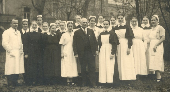 Altes Sepiafoto zeigt mittig Mann im Anzug umgeben von Diakonissen-Damen mit langer schwarzer Gewandung und weißen Hauben oder weißen Kittelschürzen. Am Rand der Gruppe steht ein zweiter Mann im Arztkittel.