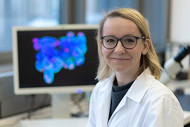 Forschung: Wie kommunizieren Darmbakterien mit dem Körper? Porträtaufnahme einer Wissenschaftlerin in weißer Laborkleidung. Im Hintergrund ein Bildschirm, auf dem ein blau eingefärbte Bild eines Darms mit einzelnen roten Punkten zu sehen ist.