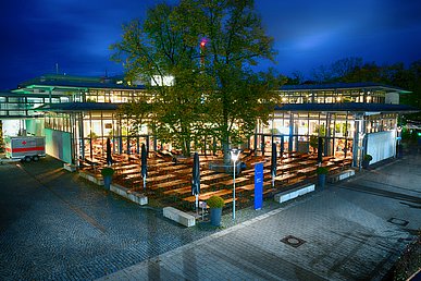 Nachtansicht der halbkreisförmigen Glasfront des erleuchteten Mitarbeiterrestaurants. Vor dem halbkreisförmigen, flachen Gebäude der Außenbericht mit Biergartengarnituren.