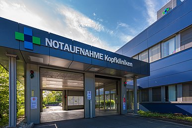 Zweiseitig befahrbare Halle mit der Beschriftung Notaufnahme Kopfkliniken. Nach rechts aus der Halle erfolgt der direkte Zugang zum Klinikgebäude, dessen Fassade mit dunkelblauen Metallelementen verkleidet ist.