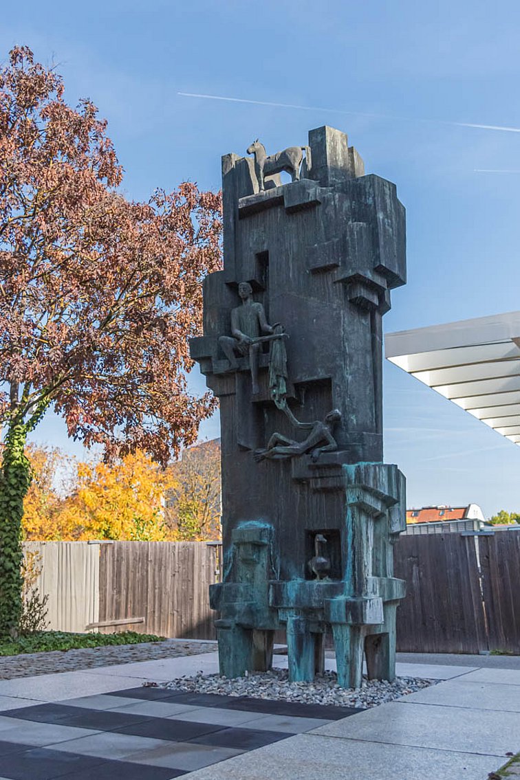 Aus ineinander übergreifenden Rechtecken bestehende längliche Skulptur die auf 6 kurzen Tischbeinen aus Stein steht. In Nischen und auf Vorsprüngen sind von oben nach unten 4 verschiedene Wesen positioniert.