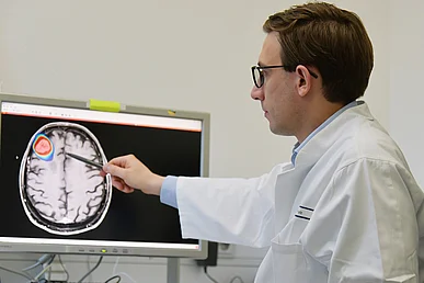 Radiomics: Tumoren mithilfe künstlicher Intelligenz genauer erkennen Mann im Arztkittel zeigt auf einem Monitor, auf dem ein Hirnscan zu sehen ist. Ein Hirnareal ist ellipsenförmig in verschiedenen Farben hervorgehoben.