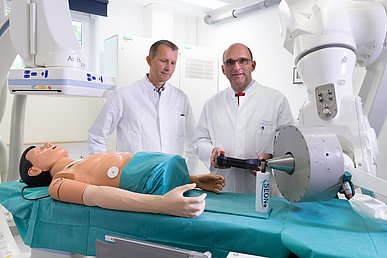 Forschung zur Nanomedizin in der Hals-Nasen-Ohren-Klinik – Kopf- und Halschirurgie Zwei Männer in weißen Kitteln stehen vor einem weißen Untersuchungsgerät mit einer Liege. Auf dem grünen Tuch auf der Liege befindet der Oberkörper einer männlichen Puppe sowie eine weiße Flasche mit der Aufschrift SEON.