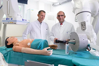 Forschung zur Nanomedizin in der Hals-Nasen-Ohren-Klinik – Kopf- und Halschirurgie Zwei Männer in weißen Kitteln stehen vor einem weißen Untersuchungsgerät mit einer Liege. Auf dem grünen Tuch auf der Liege befindet der Oberkörper einer männlichen Puppe sowie eine weiße Flasche mit der Aufschrift SEON.