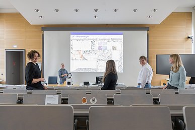 Forschungsgruppe im Pathologischen Institut Fünf Personen stehen in einem Hörsaal und blicken teils in die Kamera, teils auf eine Projektion mit Diagrammen und der Beschriftung AG Uropathologie.