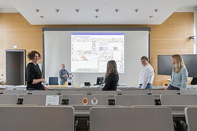 Forschungsgruppe im Pathologischen Institut Fünf Personen stehen in einem Hörsaal und blicken teils in die Kamera, teils auf eine Projektion mit Diagrammen und der Beschriftung AG Uropathologie.