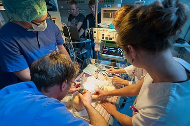 Mehrere Ärzte und Ärztinnen proben in einer Simulation die intensivmedizinische Notversorgung eines Säuglings anhand einer Puppe. Im Hintergrund werden die Daten werden zwei Mitarbeiter die Behandlungsdaten.