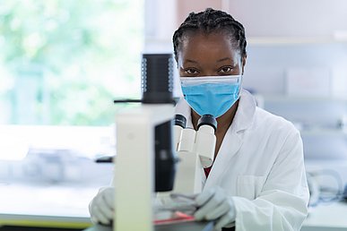 Porträtaufnahme einer jungen Frau mit weißem Kittel und blauer Schutzmaske, die von einem Mikroskop aufblickt und direkt in die Kamera schaut.
