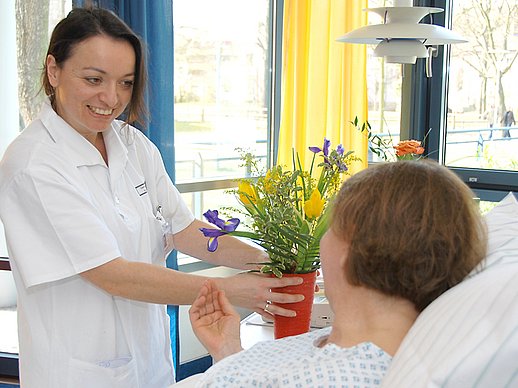 Pflegerin stellt Patientin lächelnd Blumen auf Nachttisch.