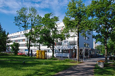Der langestreckte viergeschossige Ostflügel der Kinder und Jugendklinik im Sonnenlicht, im Vordergrund die Grünanlage des Maximiliansplatzes.